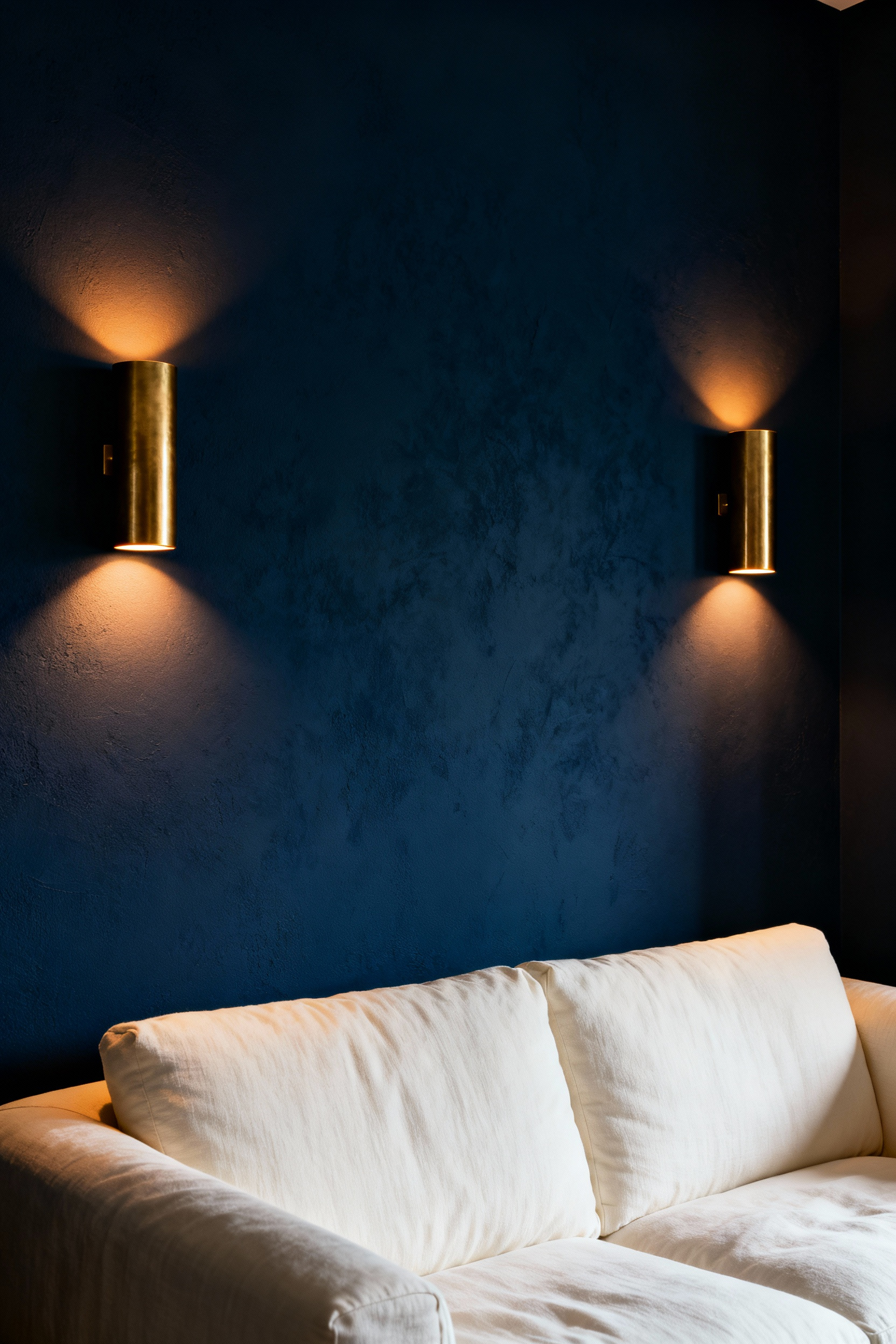 Pair of brass wall sconces providing warm, horizontal illumination in a cozy, dark blue living room, creating a soft, intimate ambiance.