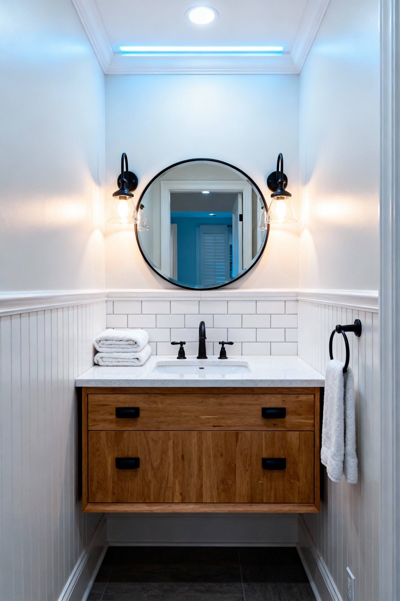 Small remodeled bathroom showing layered lighting from modern matte black sconces and recessed overhead light, complementing the new cabinet hardware on the wooden vanity.